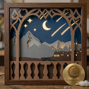 Puede incluir: Un cuadro de sombras de papel en capas en un marco de madera oscura que muestra una escena nocturna con una luna creciente, estrellas y estrellas fugaces sobre una cordillera y la silueta de una ciudad. Un medallón dorado dice Sombra & Luz Studio.