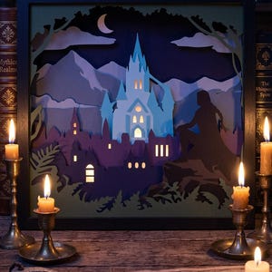 Puede incluir: Una caja de sombras de papel en capas dentro de un marco negro que muestra un castillo azul claro retroiluminado, la silueta de una ciudad morada oscura, una luna creciente y una figura oscura sentada sobre una roca ante picos montañosos.
