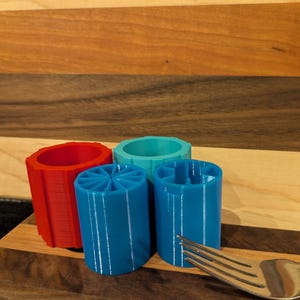 May include: A collection of four cylindrical plastic objects in red, teal, and blue, each featuring distinct internal dividers or patterns. A silver metal fork is positioned in the foreground, providing a sense of scale for these small items.
