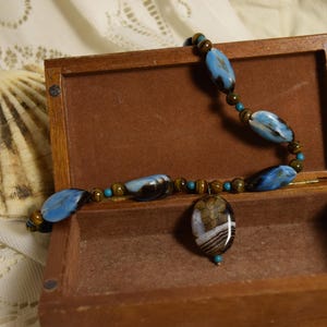 Può includere: Una collana con perline ovali marmorizzate blu e nere, piccole perline occhio di tigre marroni e minuscoli distanziatori turchesi. Il ciondolo centrale è una pietra ovale con motivi a strisce marroni, bianche e nere in una scatola.