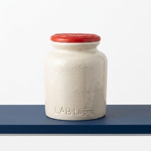 May include: A cylindrical off-white ceramic jar with a smooth finish and a bright red lid. The base of the jar features the embossed text LAB-Lagny. The container has a wide opening and a slightly tapered neck, presenting a simple, functional design.