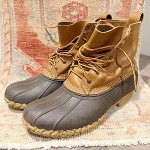 Op de afbeelding: Een paar enkelhoge duck boots met een beige leren schacht en een donkerbruine rubberen onderkant. De laarzen hebben een vetersluiting aan de voorzijde met dunne, oranjeachtige veters, metalen oogjes en een rubberen zool met textuur.