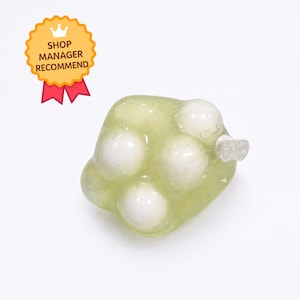 May include: A translucent light green resin bead shaped like a cluster of grapes with embedded opaque white spherical elements. The bead features a small clear loop at the side for attachment. A yellow badge with the text SHOP MANAGER RECOMMEND is visible.