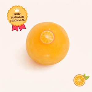 Puede incluir: Una pastilla de jabón redonda y translúcida de color naranja con un pequeño diseño de rodaja de naranja incrustado en la parte superior. Una insignia amarilla con un icono de corona y el texto SHOP MANAGER RECOMMEND está arriba a la izquierda.