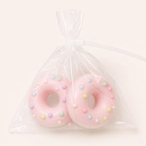 May include: Two pink glazed doughnuts with small, multicoloured round sprinkles, enclosed in a clear plastic bag that is sealed at the top with a white plastic zip tie. The doughnuts are positioned side by side within the transparent packaging.