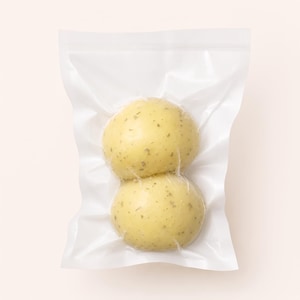 May include: Two round, pale yellow food items with small green herb flecks are sealed together inside a clear, rectangular plastic vacuum-sealed bag. The items have a smooth, firm appearance and are stacked vertically within the transparent packaging.