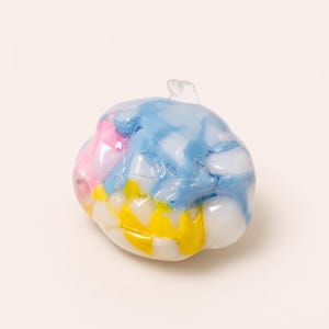 May include: A rounded glass bead featuring a multi-coloured mosaic pattern of opaque white, bright yellow, light blue, and soft pink segments. The surface has a glossy, smooth finish with a small clear glass loop at the top for attachment.