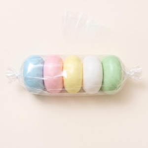 Puede incluir: Cinco objetos lisos de forma ovalada en colores pastel—azul claro, rosa, amarillo, blanco y verde claro—están dispuestos en fila y sellados dentro de un envoltorio de plástico transparente, retorcido en ambos extremos como un caramelo.