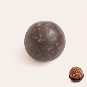 May include: A large, smooth, dark grey sphere with subtle reddish-brown and white mottled inclusions, positioned next to a small, round, textured milk chocolate confection in a brown paper liner, showcasing a contrast in size and surface finish.