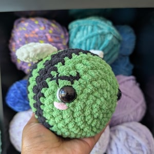 May include: A round, crocheted plush toy shaped like a bee, featuring a green and black striped body, white wings, large black plastic safety eyes, and a small pink stitched mouth. The textured yarn creates a soft, fuzzy appearance.