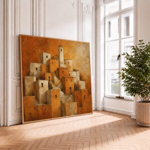 May include: A square wall art print featuring a cluster of buildings in shades of orange, terracotta, and cream. The painting depicts a hillside village with interconnected staircases and a central tower, rendered in a textured, painterly style.