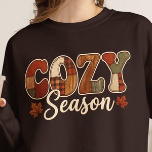 May include: A dark brown long-sleeve sweatshirt featuring the text "COZY Season" in a cream-coloured font. The word "COZY" has a multi-coloured plaid and patchwork pattern in shades of red, orange, and green. Two small maple leaves flank the text.