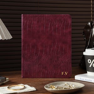 Puede incluir: Un estuche rectangular para tableta o documentos en color burdeos intenso con una textura que imita la piel de caimán. En la esquina inferior derecha presenta las iniciales F.V. en color dorado. El estuche tiene un diseño estructurado y vertical.