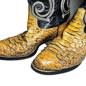 May include: A pair of cowboy boots featuring a textured, tan-colored scale pattern on the foot and a black leather upper shaft. The black shaft is decorated with a prominent, light-colored embroidered swirl design on the side of each boot.