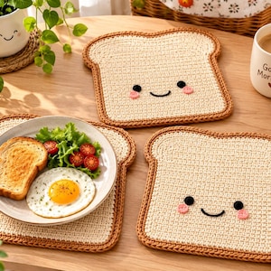 Puede incluir: Tres posavasos de ganchillo cuadrados con forma de rebanadas de pan, con centro color crema claro y borde de corteza marrón. Cada posavasos presenta una cara sonriente negra bordada con dos pequeñas mejillas rosas. Uno sostiene un plato con comida.