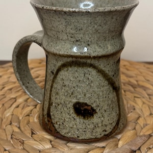 Puede incluir: Una taza de cerámica de color gris claro moteado con un borde acampanado y un asa curva. El cuerpo presenta una marca circular de color marrón oscuro con un punto central oscuro cerca de la base, con un acabado texturizado y terroso.