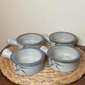 May include: A set of four round ceramic bowls with handles, featuring a speckled off-white glaze and a dark grey rim. Each bowl is decorated with a painted motif of green olives and thin grey leaves on the exterior side, showcasing a rustic finish.