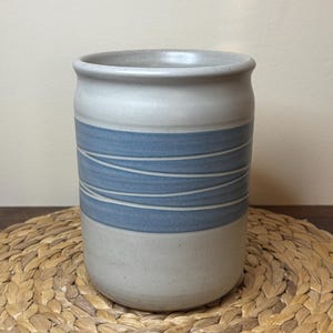 Peut inclure: Un pot en céramique cylindrique avec une finition gris clair mouchetée. L'extérieur présente une large bande horizontale de glaçure bleue mate accentuée par de fines lignes blanches ondulées non émaillées qui entourent le centre.