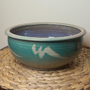 Peut inclure: Un bol en céramique rond présentant un extérieur vert et bleu sarcelle vibrant avec un motif de coups de pinceau abstraits blancs. L'intérieur du bol a un vernis violet profond et lisse, et le bord est fini avec une bande de couleur neutre.