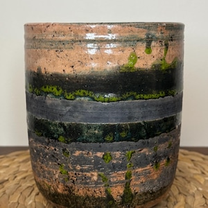 May include: A cylindrical ceramic cup featuring a textured, multi-layered glaze in shades of terracotta, charcoal black, and vibrant moss green. The surface displays horizontal bands and organic, dripping glaze patterns with a speckled finish.