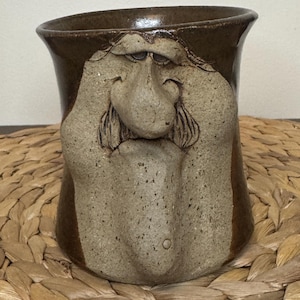 Può includere: Una tazza in ceramica con bordo e base smaltati in marrone scuro, caratterizzata da un volto scolpito color beige chiaro con un naso prominente, baffi e un mento lungo. La superficie ruvida del volto contrasta con lo smalto liscio e scuro.