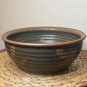 May include: A round, deep ceramic bowl with a dark green, speckled glaze and horizontal ribbed texture along the exterior. The rim features a contrasting reddish-brown tone, and the interior surface displays a smooth, dark green finish.