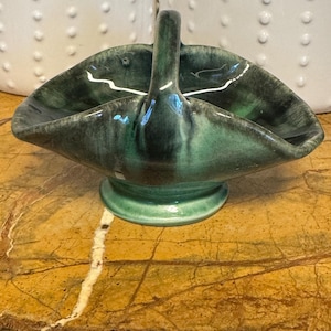 May include: A ceramic basket with a glossy finish, featuring a deep forest green and black glaze. The vessel has a central arched handle, a wide, undulating rim with four distinct points, and a circular pedestal base, creating a sculptural form.