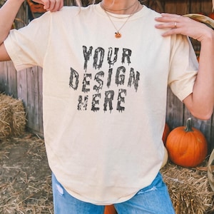 Puede incluir: Una camiseta de manga corta color crema con el texto negro "YOUR DESIGN HERE" en una fuente de estilo terror con efecto de goteo. La camiseta se combina con pantalones vaqueros azules, creando un estilo casual para eventos otoñales.