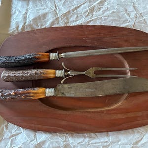 May include: A three-piece carving set featuring a knife, a two-pronged fork, and a sharpening steel. Each tool has a textured, brown antler-style handle with a silver-toned metal ferrule. The set is displayed on a dark brown wooden board.
