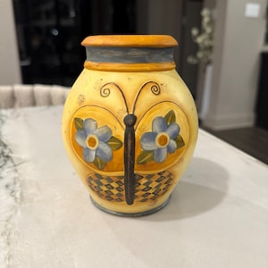 Puede incluir: Un jarrón de cerámica con base de color amarillo que presenta un diseño de mariposa pintado. La mariposa tiene flores azules en las alas superiores y un patrón de cuadros blancos y negros en las inferiores. El borde y la base tienen detalles azules.