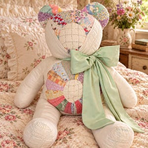 May include: A stuffed teddy bear constructed from quilted fabric featuring a patchwork pattern of various floral and geometric prints in pastel colors. The bear has a cream-colored quilted face and torso, and a large, light green satin ribbon bow.