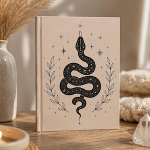 May include: A hardback book with a light beige cover featuring a black illustration of a coiled snake. The snake is decorated with celestial star patterns and is flanked by two vertical leafy branches. Small star icons surround the central design.