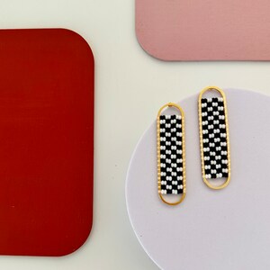 May include: A pair of gold-toned, oblong earrings. Each earring features a black and white checkered pattern within a gold frame. The earrings are displayed on a white surface, with colorful, rounded rectangular shapes in the background.