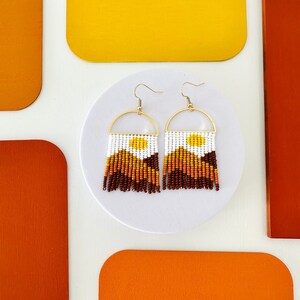 May include: A pair of beaded earrings with a gold hoop and a fringe of brown, orange, and yellow beads. The earrings have a white and yellow design in the center.