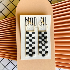 May include: Black and white checkered beaded earrings with gold-colored hooks. The earrings are rectangular and hang from a gold-colored hoop. The earrings are displayed on a white card with the text "MODISH HANDMADE".