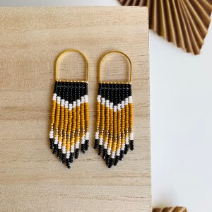 May include: A pair of beaded earrings with a gold-colored arched top and a fringe design. The earrings feature a pattern of black, white, and yellow beads. The earrings are displayed on a light wooden surface.