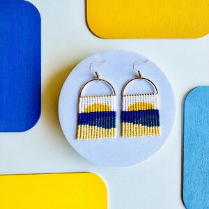 May include: A pair of beaded earrings with a white, yellow, and blue color scheme. The earrings are shaped like a sun setting over the ocean.