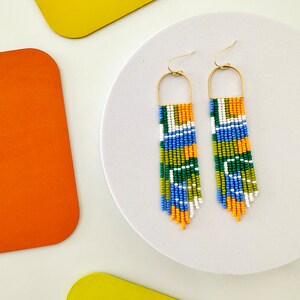 May include: A pair of long, beaded earrings with a geometric pattern in shades of blue, green, orange, and white. The earrings are suspended from gold hoops.