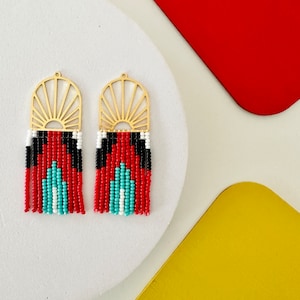 May include: A pair of gold-colored metal earrings with a sunburst design at the top. The earrings feature a beaded pattern in red, black, white, and turquoise, with a fringe of beads at the bottom. The earrings are displayed on a white surface with red and yellow geometric shapes in the background.