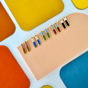 May include: A row of colorful beaded earrings with gold hooks. The earrings are arranged on a light brown surface with yellow, orange, and blue squares in the background.