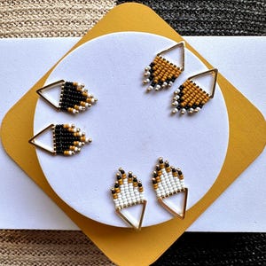May include: Three pairs of triangular earrings featuring gold-tone metal frames and small seed beads. One pair is black with white and gold accents, one is mustard yellow with black and white, and one is white with mustard yellow and black beads.