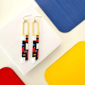May include: A pair of beaded earrings with gold-colored arched frames. The earrings feature a geometric pattern of red, black, white, and blue beads. The earrings are displayed on a white surface with colorful geometric shapes in the background.