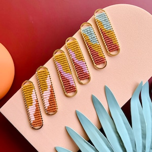 May include: Six beaded bar earrings in a variety of colors, including yellow, pink, orange, and blue. The earrings are set in gold-colored frames. The earrings are arranged on a pink surface with a dark red background and a light green leaf.