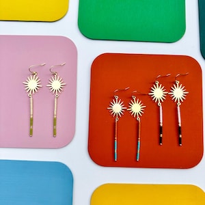 May include: Gold-tone sunburst earrings with beaded fringe. The earrings feature a sunburst design at the top and a vertical fringe of colorful beads. The earrings are displayed on pink and orange backgrounds.