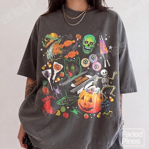 May include: A charcoal grey short-sleeve t-shirt featuring a vibrant Halloween-themed graphic print. The design includes a witch, a green skull, a black cat, a skeleton, a jack-o'-lantern candy bucket, eyeballs, candy corn, and a lava lamp.