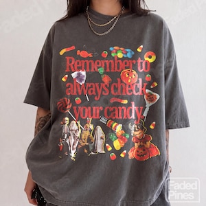 Puede incluir: Una camiseta gris carbón de manga corta con el texto en rojo "Remember to always check your candy." El gráfico incluye ilustraciones coloridas de varios dulces, un esqueleto, un fantasma y un niño con un disfraz de calabaza.