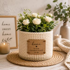 Könnte beinhalten: Ein cremefarbener gehäkelter Korb mit weißen Rosen. Auf der Vorderseite ist ein rechteckiges Holzschild angebracht, das den eingravierten Text: home, ein kleines Herzsymbol und den Satz IS OUR FAVORITE PLACE TO BE zeigt.