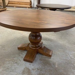 May include: A round wooden dining table with a pedestal base. The table top is a rich brown color and the base is a slightly darker brown. The table is in a workshop setting.