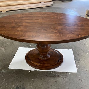May include: A round, dark brown wooden dining table. The table has a pedestal base with a bulbous center and a circular base. The tabletop is a solid, smooth surface. The table is sitting on a white surface.