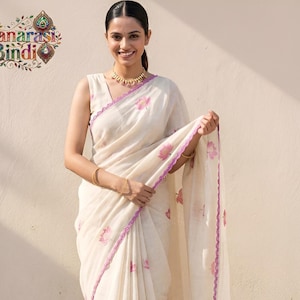 Könnte beinhalten: Ein ärmelloser Sari in Off-White mit einem sich wiederholenden Muster aus kleinen rosa Lotusblüten. Das Kleidungsstück ist an den Rändern mit einer violetten, gewellten Borte versehen. Das Logo von Banarasi Bindi ist oben links zu sehen.